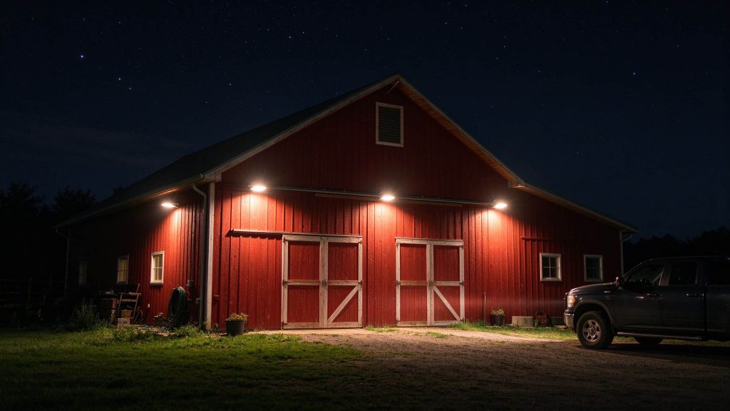 A red wooden barn at night with warm solar barn lights lit under the eaves and a pickup truck parked nearby